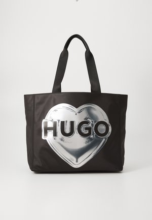 Czarna torba typu tote z czarnymi uchwytami, z dużym srebrnym sercem i napisem „HUGO” pogrubioną czcionką.
