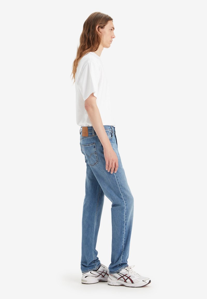 Jeans de mezclilla azul claro con corte recto, que cuentan con un parche de cuero en la parte trasera de la cintura y bolsillos con costuras prominentes. Llevados con una camiseta blanca.