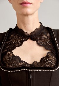 Gros plan sur une femme portant un col en dentelle noire avec un petit bouton en perle et un vêtement noir avec un bord perlée le long de l'encolure.