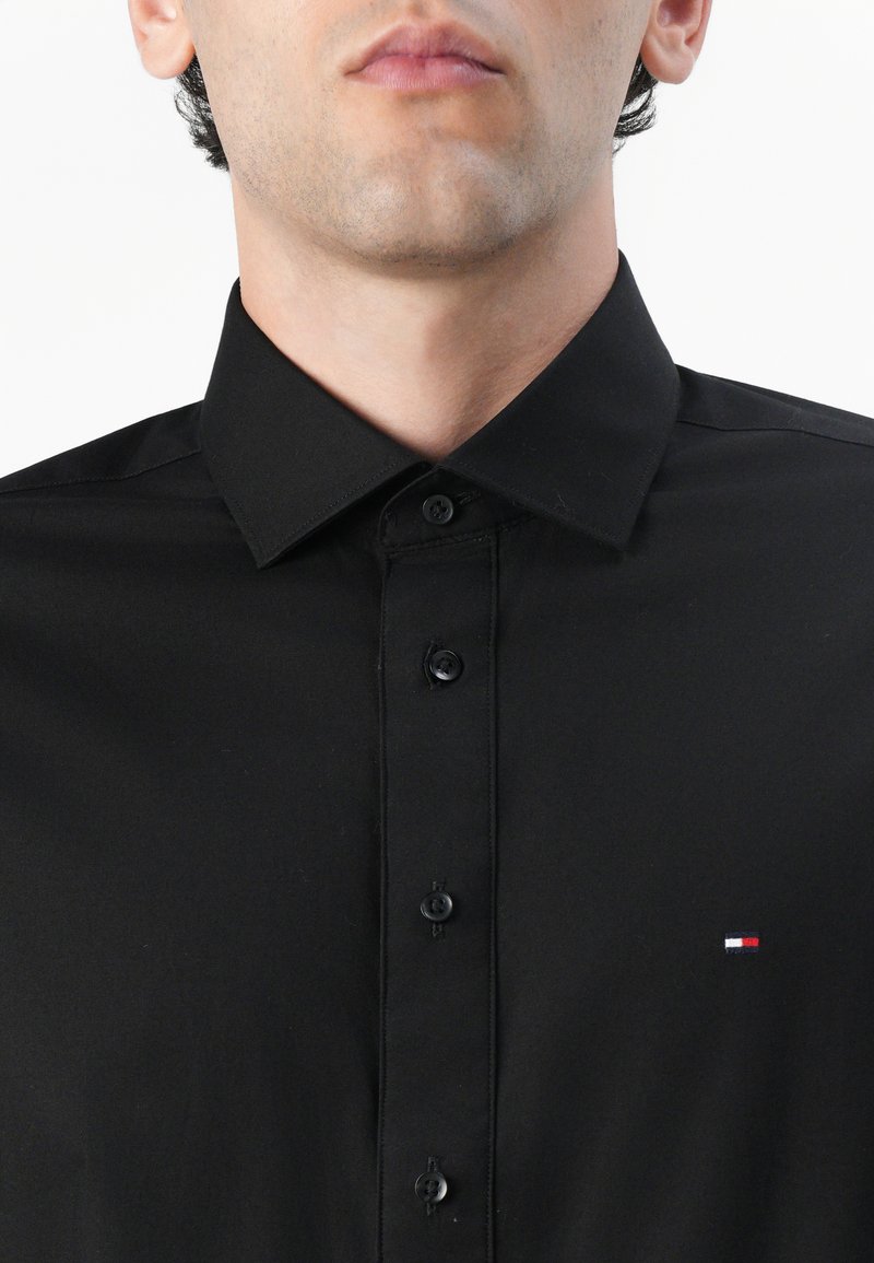 Black button-up shirt with a classic collar, featuring a small embroidered logo in red, white, and blue on the left chest.