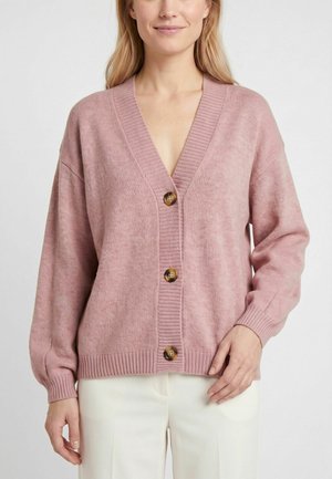 Femme portant un cardigan rose doux avec trois boutons marron sur un pantalon blanc, debout devant un fond clair uni.