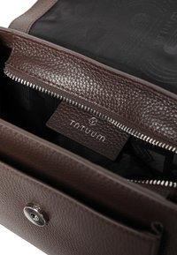 Brązowa skórzana torebka z teksturowanym wykończeniem, posiadająca zamek błyskawiczny, materiałową podszewkę oraz widoczną w środku metkę z logo.