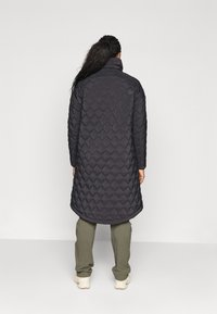 Padded coat in dark gray with a diamond quilted pattern, high collar, and long length, paired with olive green pants.