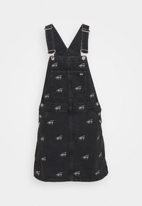 Svart denimklänning med hängslen och mönster av uniformslogotyper. Har justerbara axelband, ficka fram och detaljer i metall.