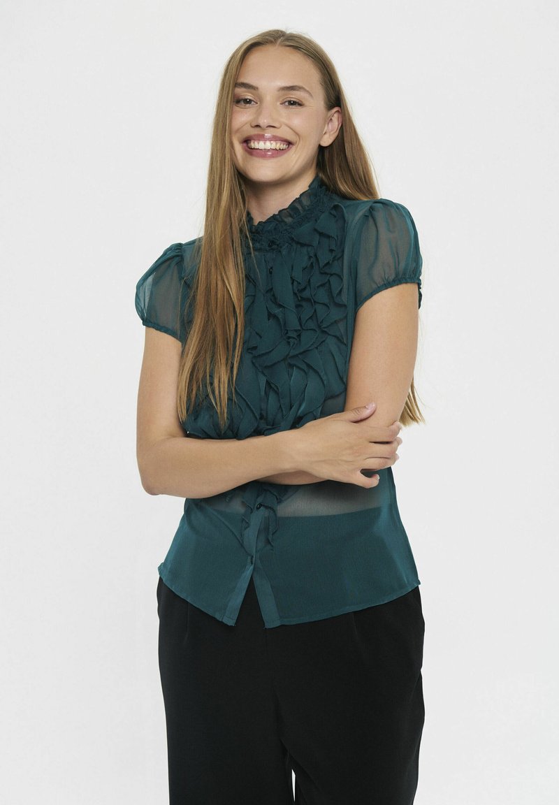 Teal ruffled blouse with short sleeves, high collar, and button closure; sheer texture with layered fabric details; paired with black pants.