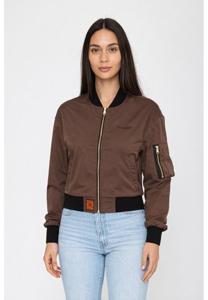 Donna che indossa una giacca bomber marrone con polsini e fascia neri, tasche con cerniera sulla manica, abbinata a jeans azzurri chiari su sfondo bianco.