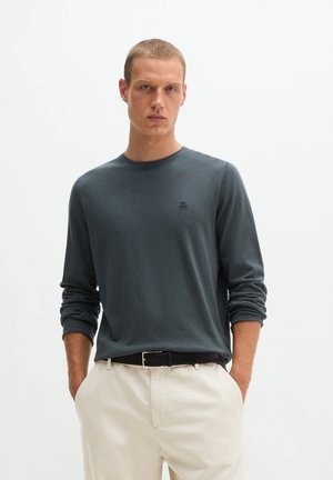 Junger Mann mit kurzen Haaren, der ein dunkelgraues Langarmshirt und hellbeige Hosen trägt, mit den Händen in den Taschen vor einem schlichten Hintergrund.