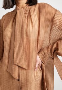 Donna che indossa una blusa leggera di colore marrone chiaro con una grande cravatta annodata al collo e la mano che riposa nella tasca laterale.