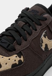 Zapatillas de ante marrón con cordones negros, que presentan un diseño de manchas en tonos beige y negro en el lateral y una suela oscura y texturizada.