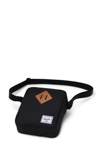 Černá látková crossbody taška s hladkou texturou, která má na přední straně hnědou koženou diamantovou aplikaci a bílou značkovou etiketu.