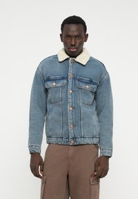 Denim striukė, pasižyminti šviesiai mėlyna spalva, avikailio apykakle, vario mygtukais, priekinėmis kišenėmis ir laisvu siluetu.