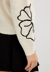 Suéter de punto en color crema con bordado floral negro en la manga izquierda, con puños acanalados y textura suave. Falda negra parcialmente visible.