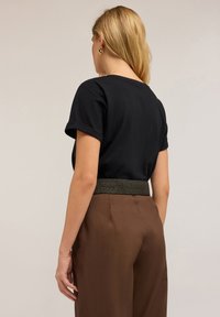 Mujer con cabello rubio que lleva pendientes de aro dorados, una camiseta negra de manga corta y pantalones marrones de tiro alto con un cinturón texturizado, de espaldas.