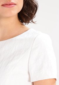 Top blanco texturizado con cuello redondo y mangas cortas, que presenta patrones geométricos sutiles y un acabado suave.