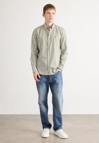 Camicia a quadri verde oliva e bianca con colletto, abbinata a jeans blu e sneaker bianchi; in piedi su uno sfondo neutro.