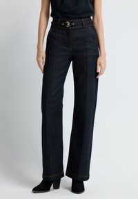 Tmavé džínové široké kalhoty mají vysoký pas, viditelné švy a pásek se zlatou sponou. Lemovky jsou mírně rozšířené.