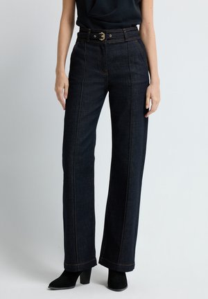 Mörk denim byxor med vida ben har hög midja, synlig sömnad och ett bälte med ett guldspänne. Bensluten är något utsvängda.