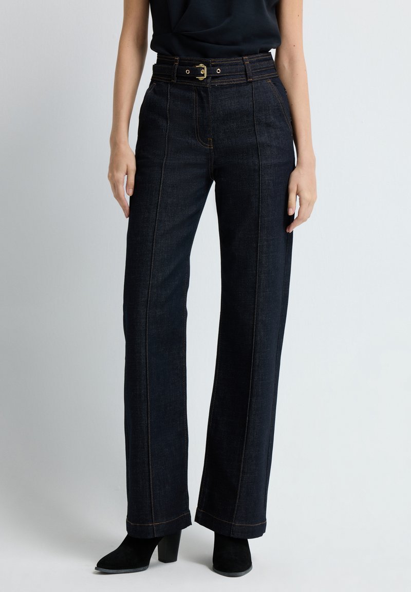 Tmavé džínové široké kalhoty mají vysoký pas, viditelné švy a pásek se zlatou sponou. Lemovky jsou mírně rozšířené.