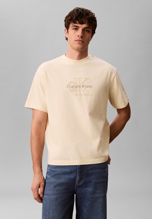 Młody mężczyzna ubrany w beżową koszulkę z logo Calvin Klein i niebieskie dżinsy, stojący na tle jednolitego szarego tła.