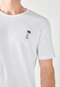 Witte katoenen T-shirt met een ronde halslijn, voorzien van een klein zwart geborduurd ontwerp van een palmboom en surfplank op de linkerborst.