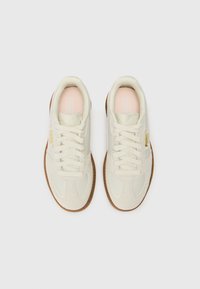 Valged sneakers, millel on nubuk- ja sünteetiline pealis, ümar varvas, tekstureeritud tald, pehmed roosad sisetallad ja paelad. Omab kuldseid brändinguelemente.