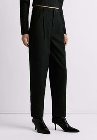 Zwarte getextureerde broek met plooien, hoge taille en zijzakken, gecombineerd met spitse zwarte hakken. Glad stof met ingewikkelde details.