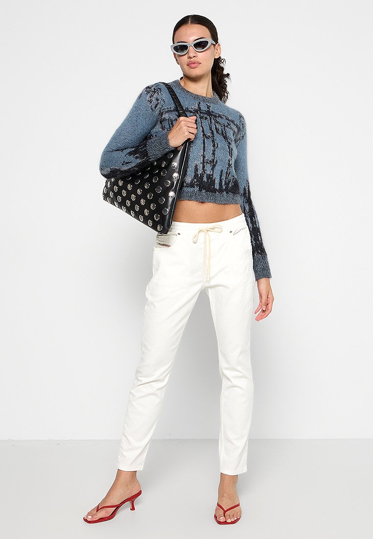 Blauer Strickpullover mit grafischem Muster, weiße schmal geschnittene Hose, rote Absatzsandalen und eine schwarze Tasche mit silbernen Akzenten.