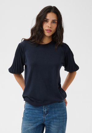 Joven mujer con cabello oscuro y ondulado, con una camiseta azul marino con mangas cortas plisadas y vaqueros azules sobre un fondo liso.