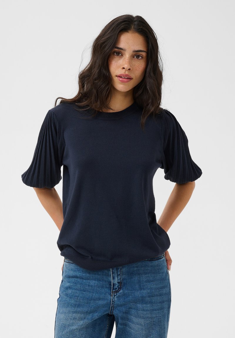 Jeune femme aux cheveux ondulés foncés, portant un haut bleu marine avec des manches courtes plissées et un jean bleu sur un fond uni.