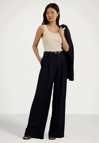 Mulher a usar um top de malha em creme, calças largas pretas com cinto, sapatos de salto pretos, segurando um blaser preto sobre um ombro.