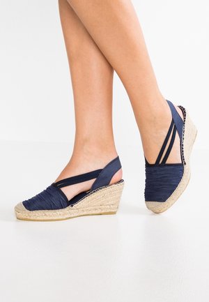 Marineblå kilehæle med overdel i tekstureret stof, juteindpakket hæl og krydsede ankelremme. Åben bagdesign, der fremhæver foden.