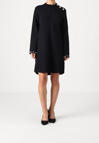 Svart långärmad klänning med pärlknappar på axlarna och ärmarna, bärs med svarta spetsiga klackskor, stående mot vit bakgrund.