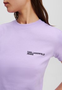 Fialové tričko z bavlneného zmesi, s okrúhlym výstrihom a krátkymi rukávmi. Na ľavej hrudi sa nachádza čierne logo "KARL LAGERFELD JEANS". Hladká textúra.
