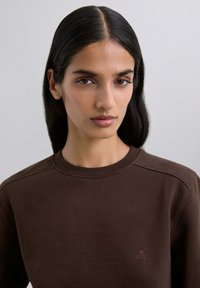 Sudadera marrón con cuello redondo y logotipo bordado en el lado izquierdo del pecho. La tela parece suave, con una textura lisa y un ajuste holgado.