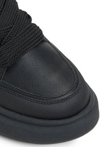 Bout de basket noir en cuir texturé avec des lacets épais tissés sur une semelle en caoutchouc épaisse, présenté sur un fond blanc.