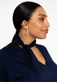 Zilveren oorhangers met een kristallen versiering, gedragen met een marineblauwe blouse met een gestrikt nekteken en een v-hals.