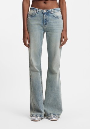 Oseba, ki nosi svetlo modre kavbojke z visokim pasom in razširjenimi hlačnicami ter srebrne superge, stoji pred belim ozadjem.