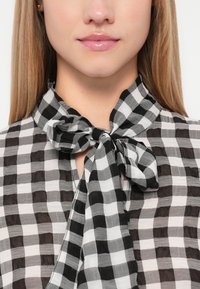 Melnbalts čekošanas blūze ar kakla saiti, izgatavota no viegla, puscaura auduma un ar atvērtu dekoltē dizainu.