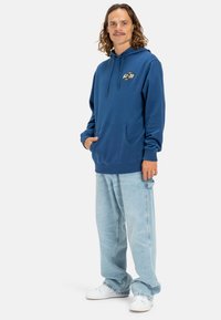 Blue hooded sweatshirt with a kangaroo pocket and logo on the chest, paired with light blue loose-fitting jeans and white sneakers.