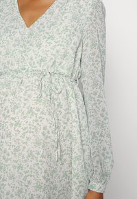Vestido de tecido leve com estampado floral, apresentando um decote em V, mangas longas bufantes e um laço na cintura. Cores verde e branco.
