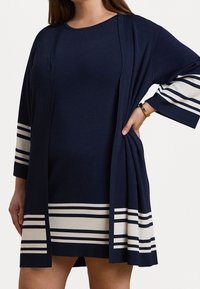 Lahti sininen mekko leveillä hihoilla, jossa on valkoisia va Horizontal-raitoja. Kangas näyttää sileältä, rento istuvuus ja avoin etusuunnittelu.