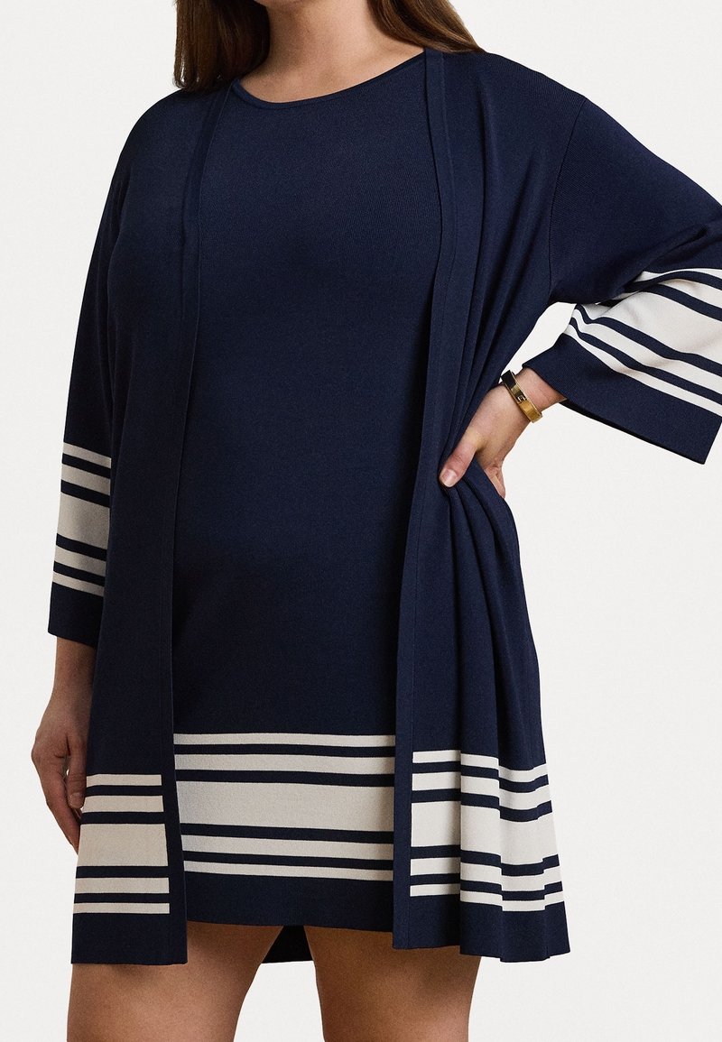 Lahti sininen mekko leveillä hihoilla, jossa on valkoisia va Horizontal-raitoja. Kangas näyttää sileältä, rento istuvuus ja avoin etusuunnittelu.