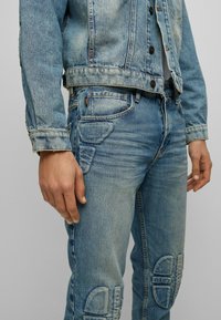 Ljusblå denimjacka och jeans har en sliten textur, kontrasterande sömmar och unika lappar på knäna.