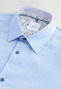 Himmelblaues, strukturiertes Hemd mit Kragen, das ein floral gemustertes Innenfutter und zwei dunkle Knöpfe auf der Vorderseite hat. Das Markenlabel ist sichtbar.