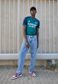 Maglia da calcio verde e blu navy con il testo "Emirates Fly Better", jeans a gamba larga azzurri chiari e sneaker bianche con dettagli rossi, in piedi contro un muro.