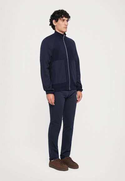 Jachetă bleumarin cu fermoar, confecționată dintr-un material fleece, având două buzunare frontale. Asortată cu pantaloni bleumarin și adidași din suede călcată. Croi lejer.
