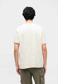 Krémové tričko s krátkým rukávem a kulatým výstřihem, vyrobené z měkké bavlny. Zadní strana je jednoduchá a má volný střih, doplněné olivově zelenými kalhotami.