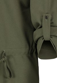 Olivgrön blus i mjukt, texturerat tyg; har uppvikta ärmar med knapppynt och dragsko i midjan för justerbar passform.