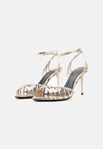 Χρυσά σανδάλια με λουράκια, ψηλό τακούνι, λουράκια στον αστράγαλο και ανοιχτά δάχτυλα, σε απλό λευκό φόντο.