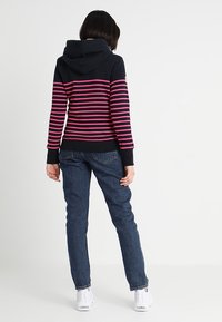 Felpa con cappuccio blu scuro con strisce rosa, texture morbida, polsini e orlo a costine. Abbinata a jeans blu scuro e sneakers bianche.